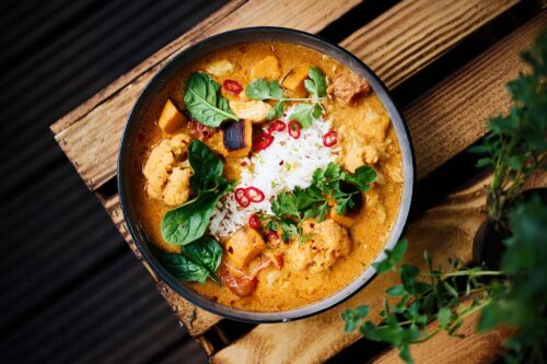 Draufsicht auf ein frisch zubereitetes Curry-Gericht mit frischen Kräutern auf einem rustikalen Holztisch.